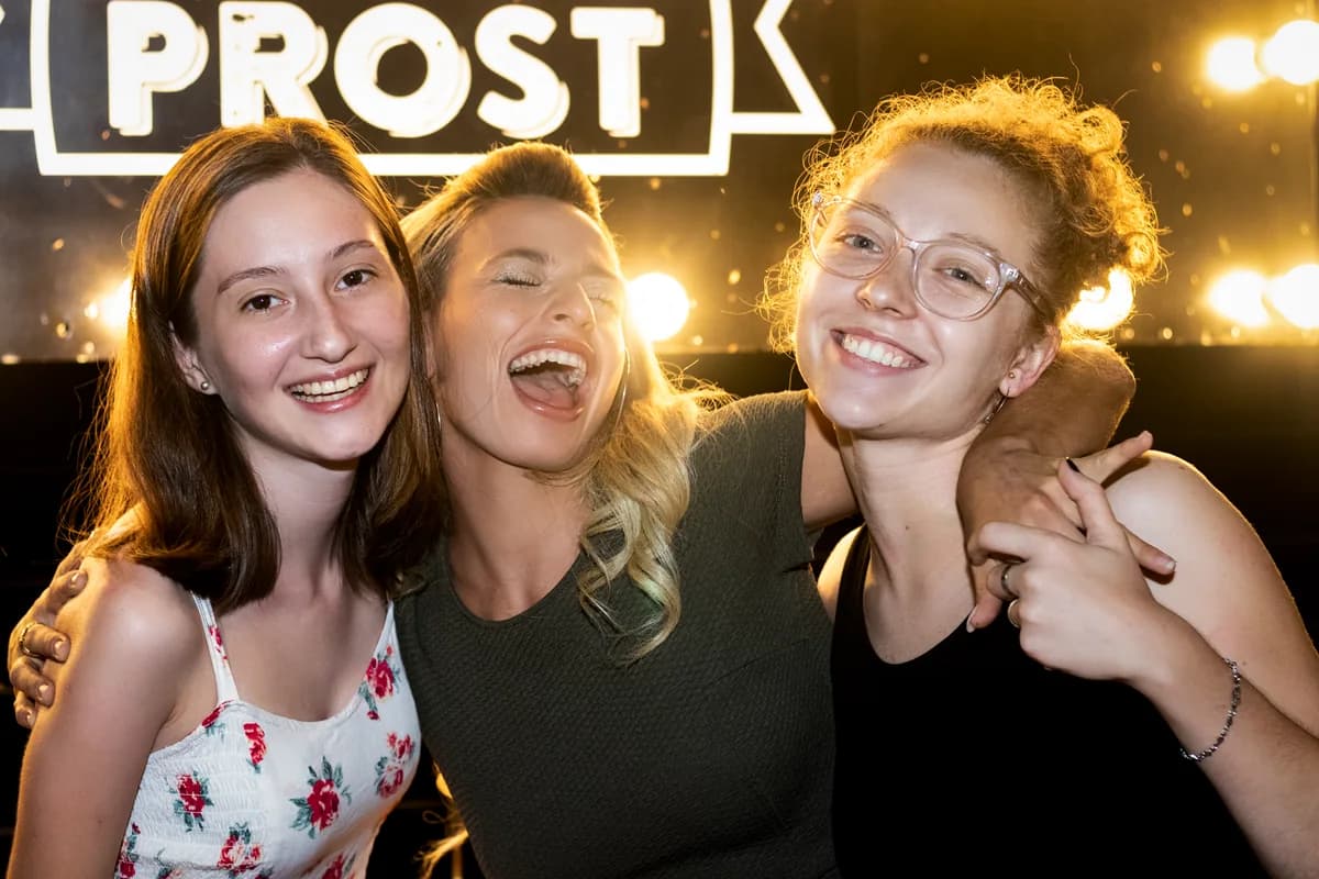 Amigas sonriendo bajo el letrero luminoso de Prost